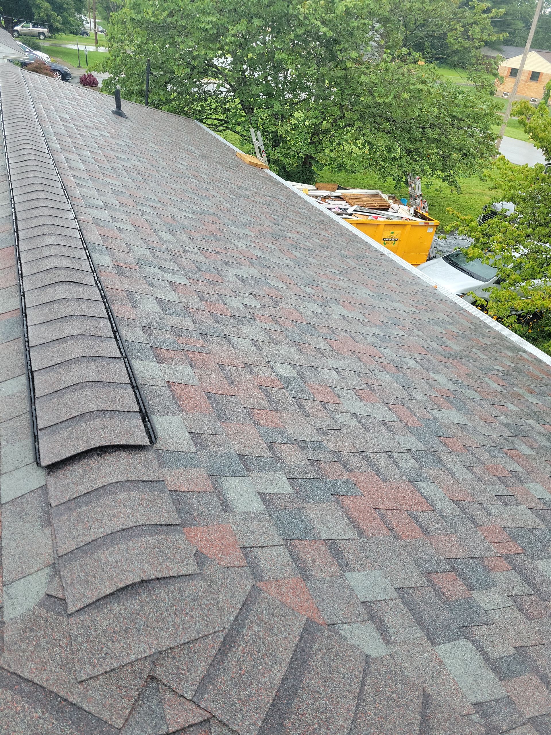 A high-angle view of a newly shingled roof with a ridge vent, seen from above with a yellow dumpster in the background.
