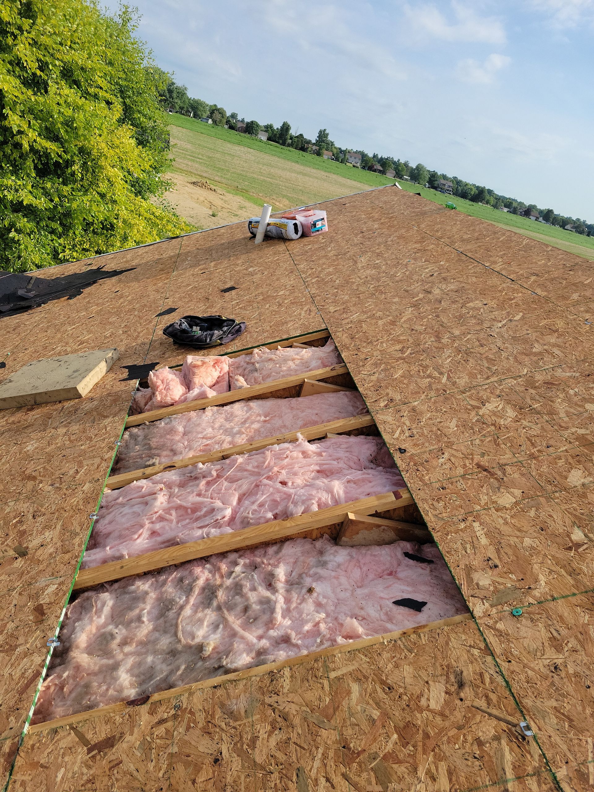 A rectangular hole cut into a roof exposing the wooden rafters and pink insulation underneath, with a field in the back.