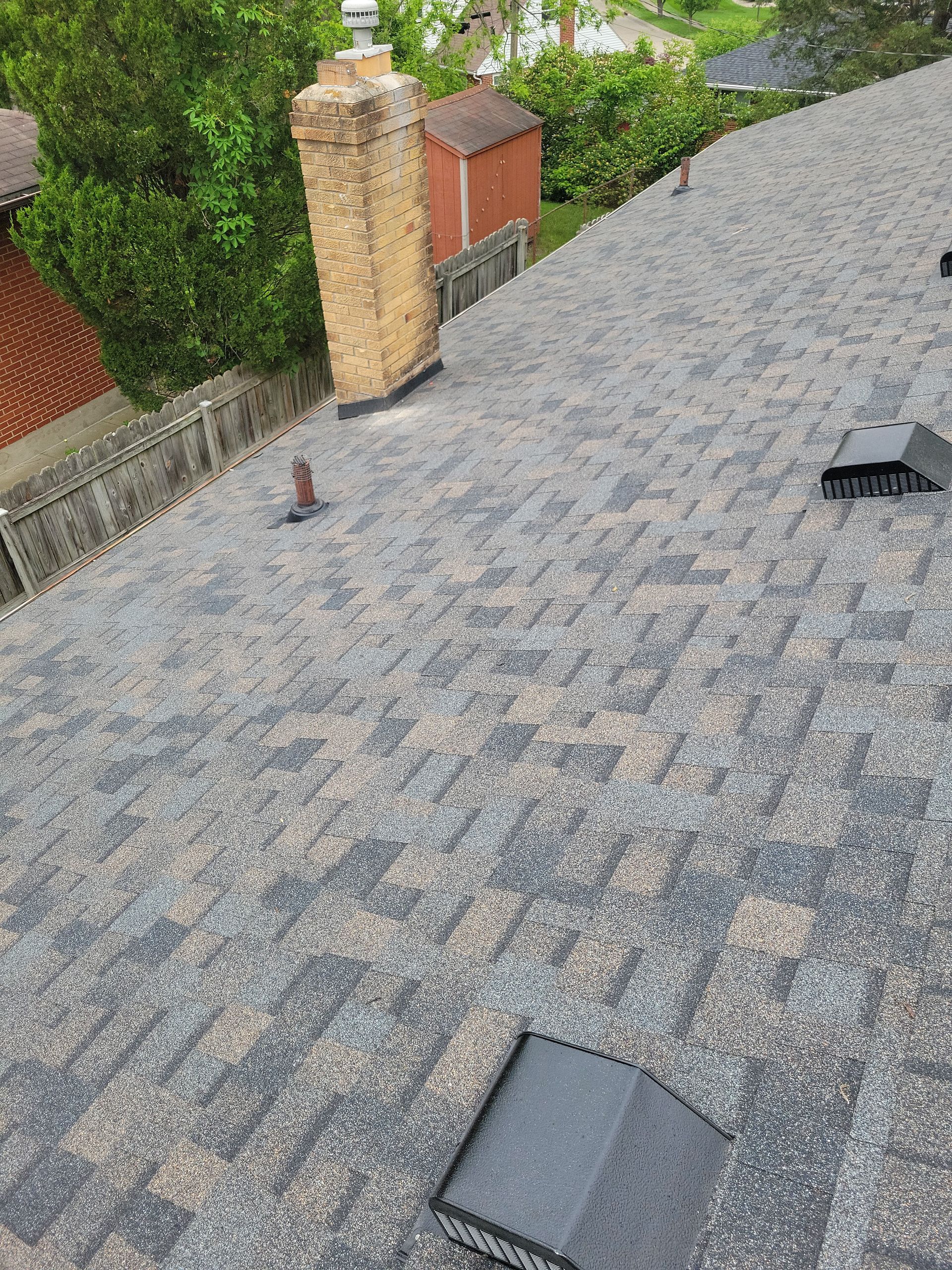 A steep roof covered in grey and brown asphalt shingles, featuring a stone chimney and several black vents.