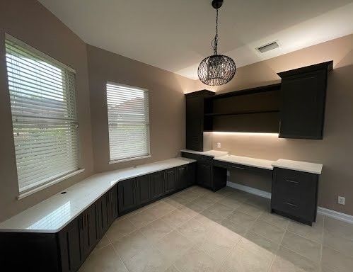 Empty home office with built-in dark cabinets, white countertops, and two windows with blinds.