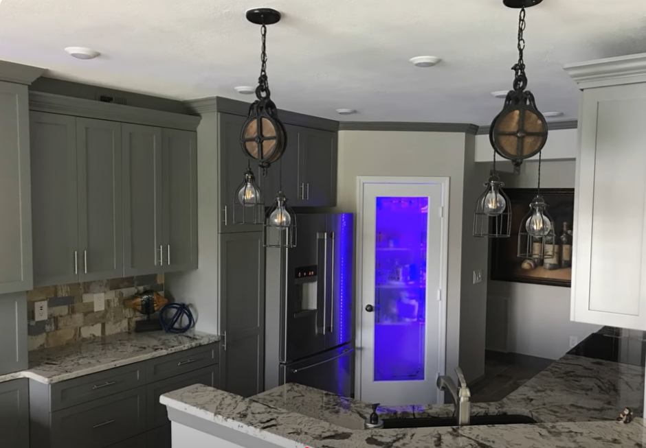 Modern kitchen with gray cabinets, granite island, and hanging pendant lights; purple-lit glass door in back.