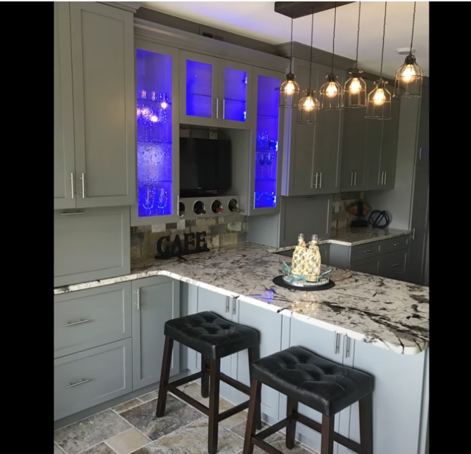 Modern kitchen with gray cabinets, blue-lit glass cabinet, marble island, and two black stools