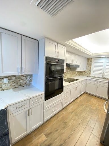 Bright white galley kitchen with wood floors, built-in oven, white cabinets, and stone backsplash.