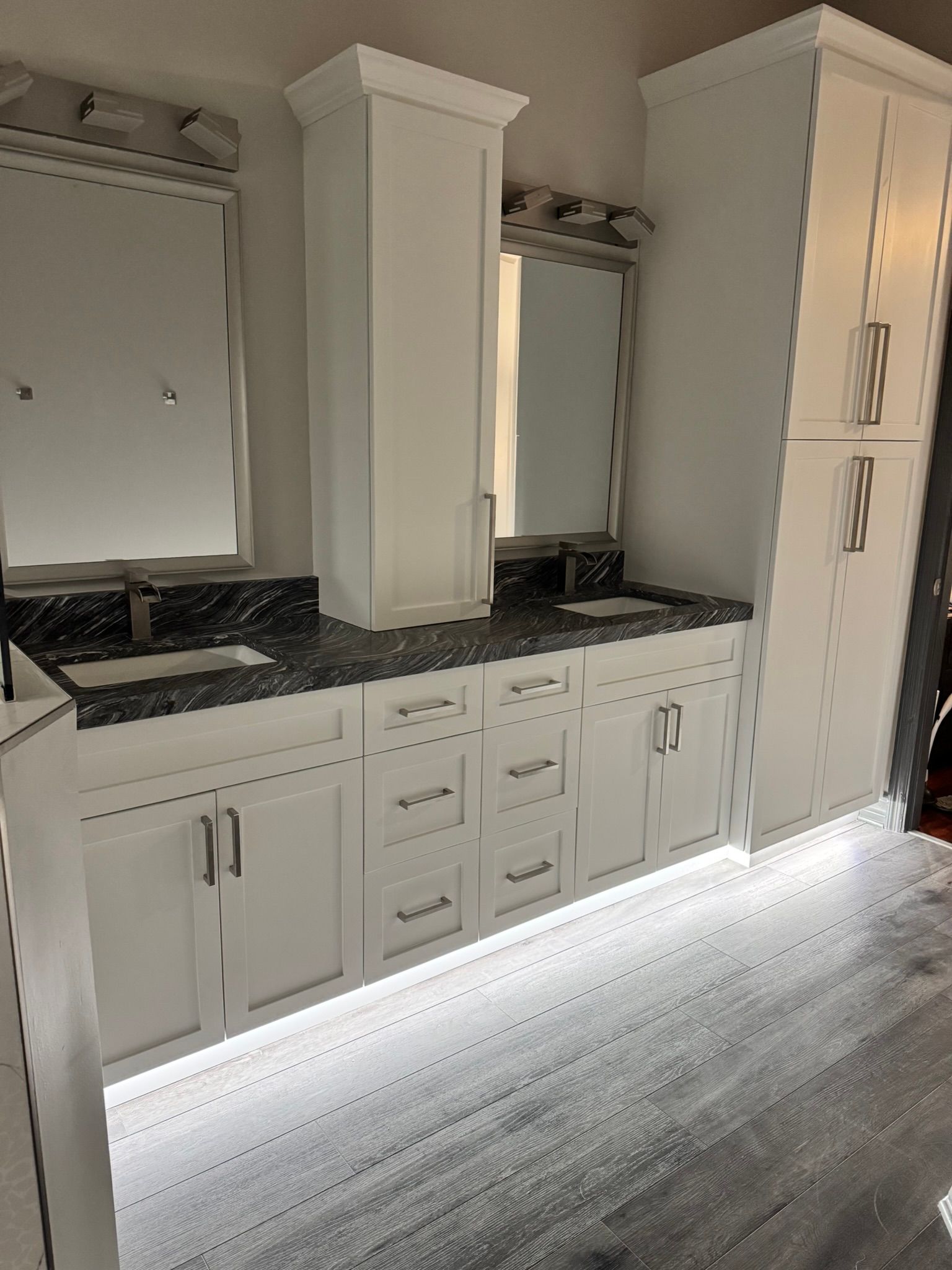 Bright white bathroom vanity with double sinks, mirrors, dark countertop, and tall cabinets along a wood floor