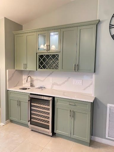 Built-in sage green bar cabinet with wine fridge, countertop, and glass-front storage in a bright room