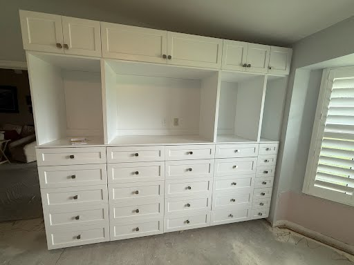 White built-in dresser and shelving wall in a bedroom with a slanted ceiling and window shutters.