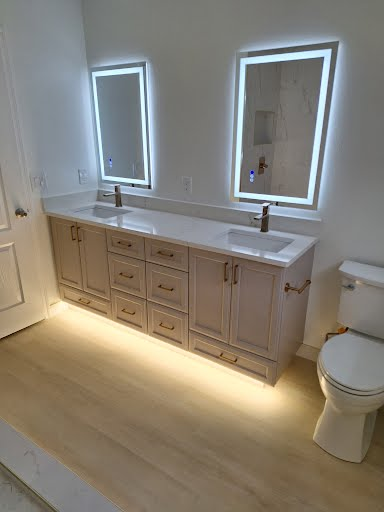 Bright bathroom vanity with two illuminated mirrors, beige cabinets, white countertop, and a toilet at right.