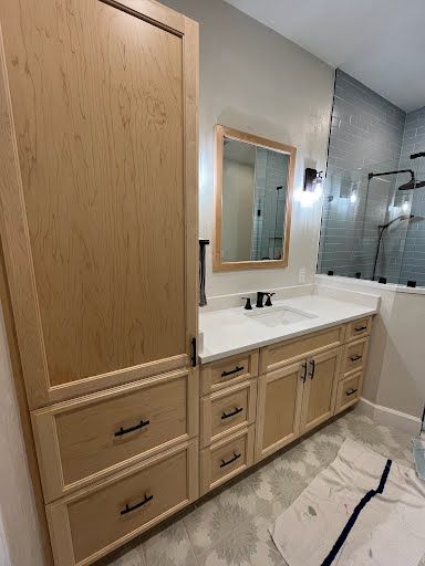 Modern bathroom vanity with light wood cabinets, double sink, mirror, and white tub/shower area
