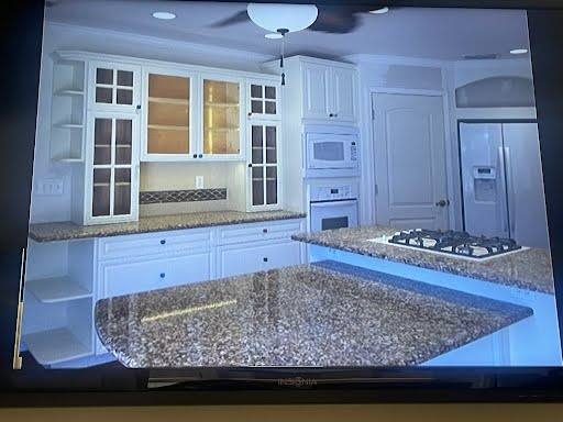 Bright white kitchen with granite island, gas stove, built-in oven, cabinets, and stainless steel refrigerator.
