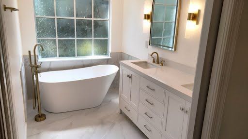 Bright modern bathroom with freestanding tub, vanity, large window, and wall sconces.