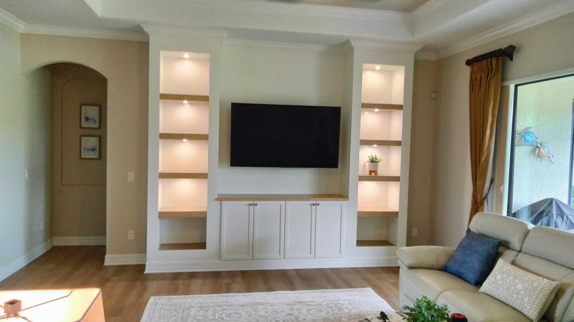 Bright living room with wall-mounted TV, built-in shelves, beige walls, and a sofa by a window