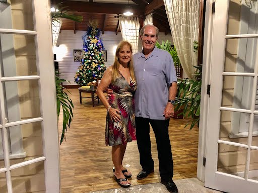 Two people smiling in a doorway decorated with lights and a Christmas tree in the background.