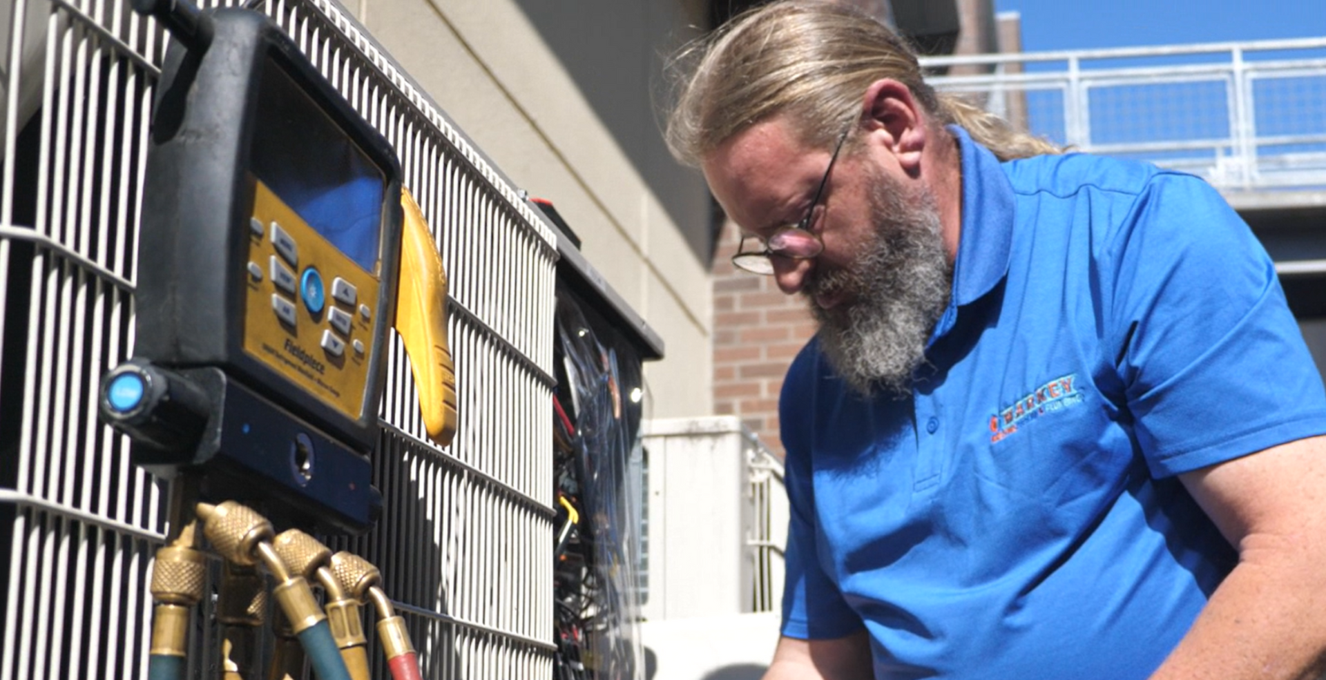 HVAC technician servicing an air conditioning unit. He's wearing a blue shirt and using a gauge tool.