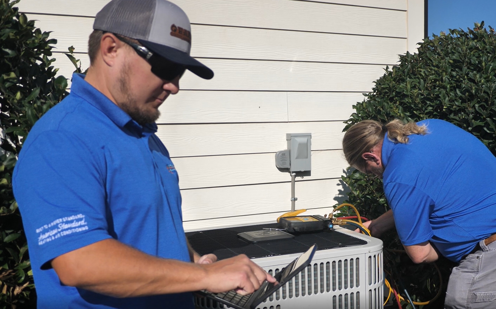 Two HVAC technicians in blue shirts working on an air conditioning unit outside. One uses a laptop.