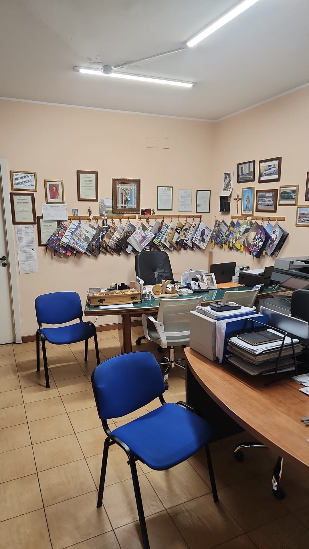 Ufficio con sedie blu, scrivanie, documenti, foto incorniciate e una parete con opere d'arte o oggetti artigianali appesi.
