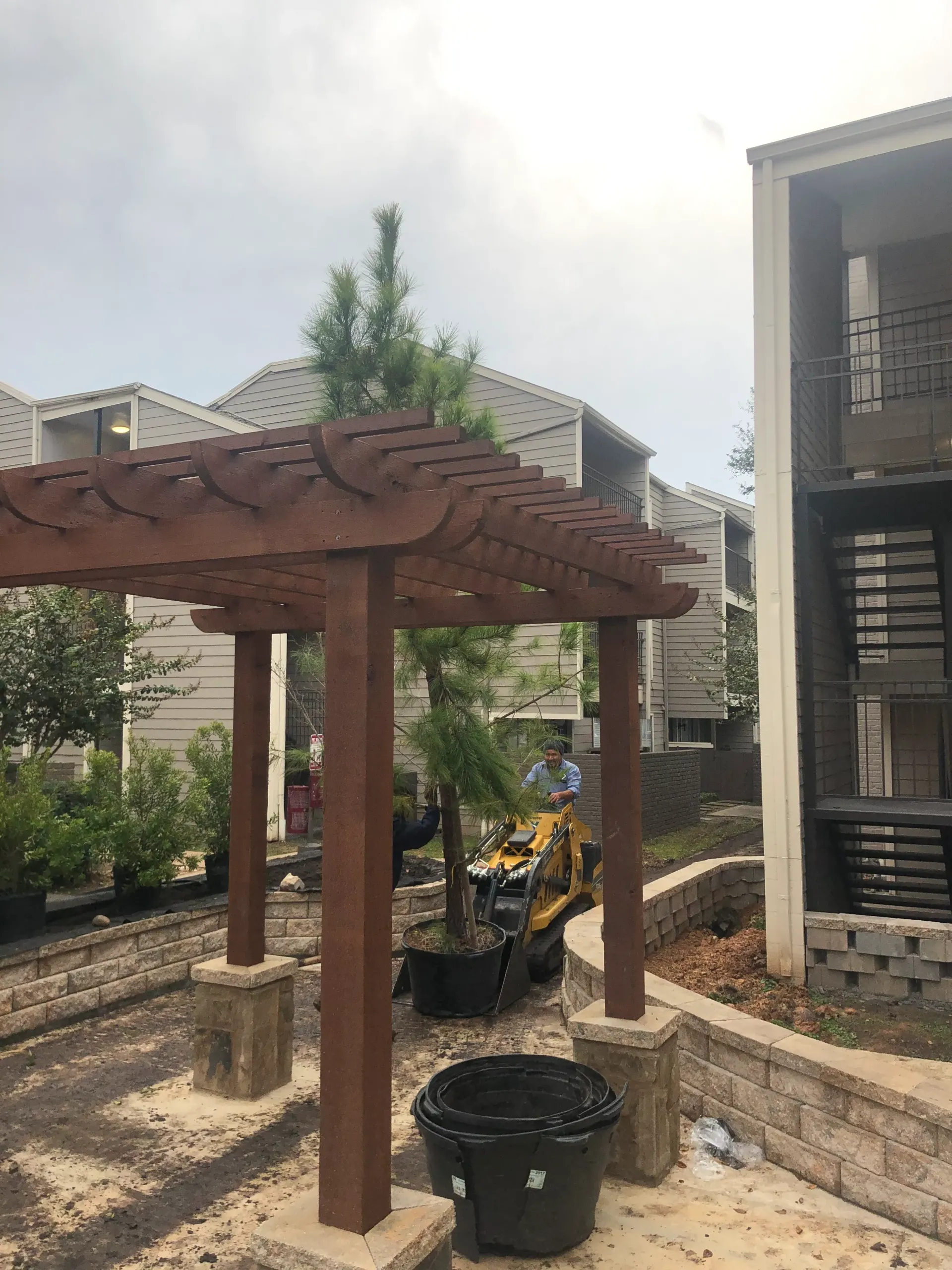 Rustic wooden pergola in a landscaped courtyard beside a stone building, with construction equipment in back.