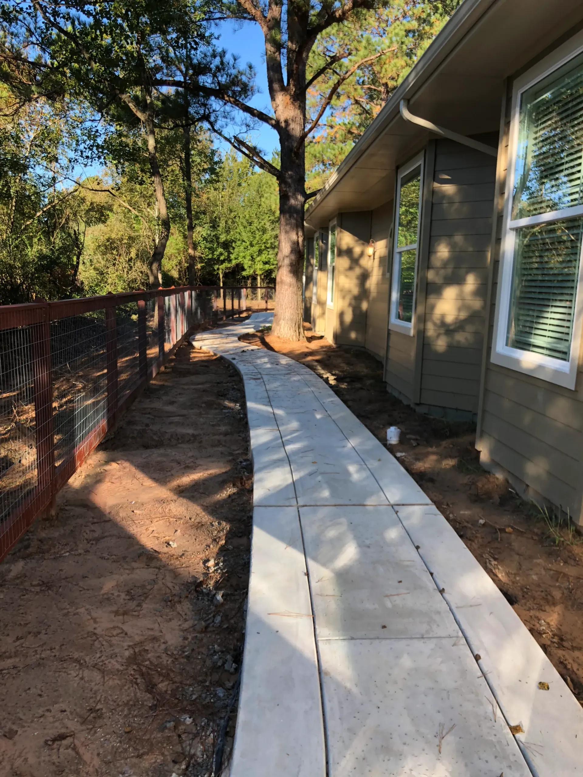 Sidewalk beside a house, with a fenced yard and trees along the path.