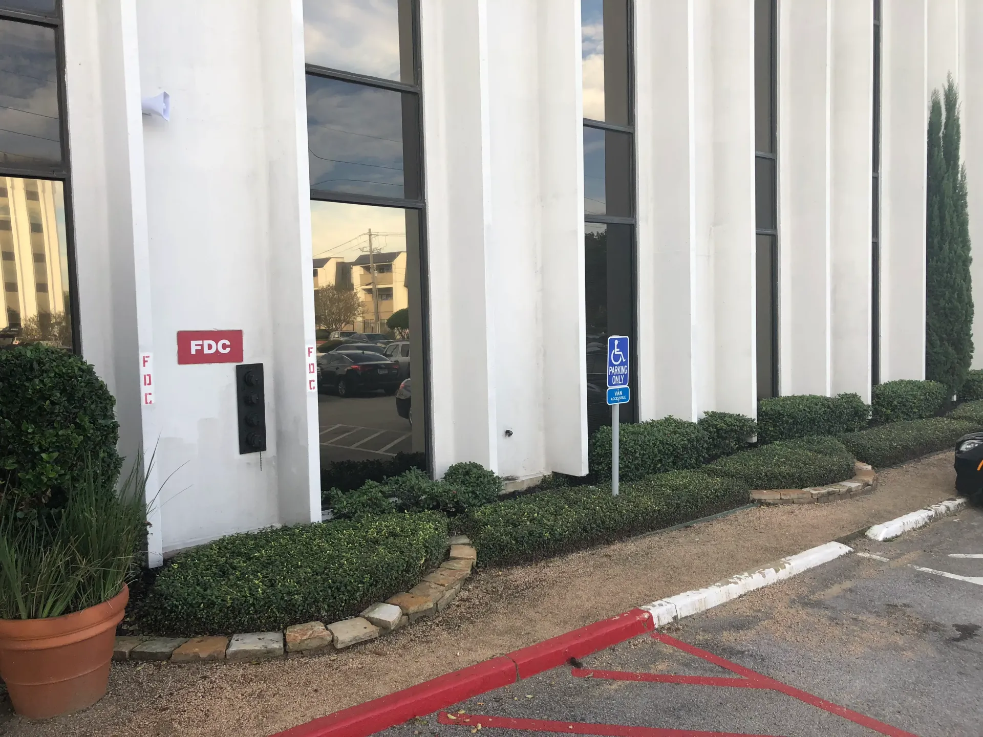 Modern office building entrance with white columns, reflective windows, red curb, and landscaped shrubs