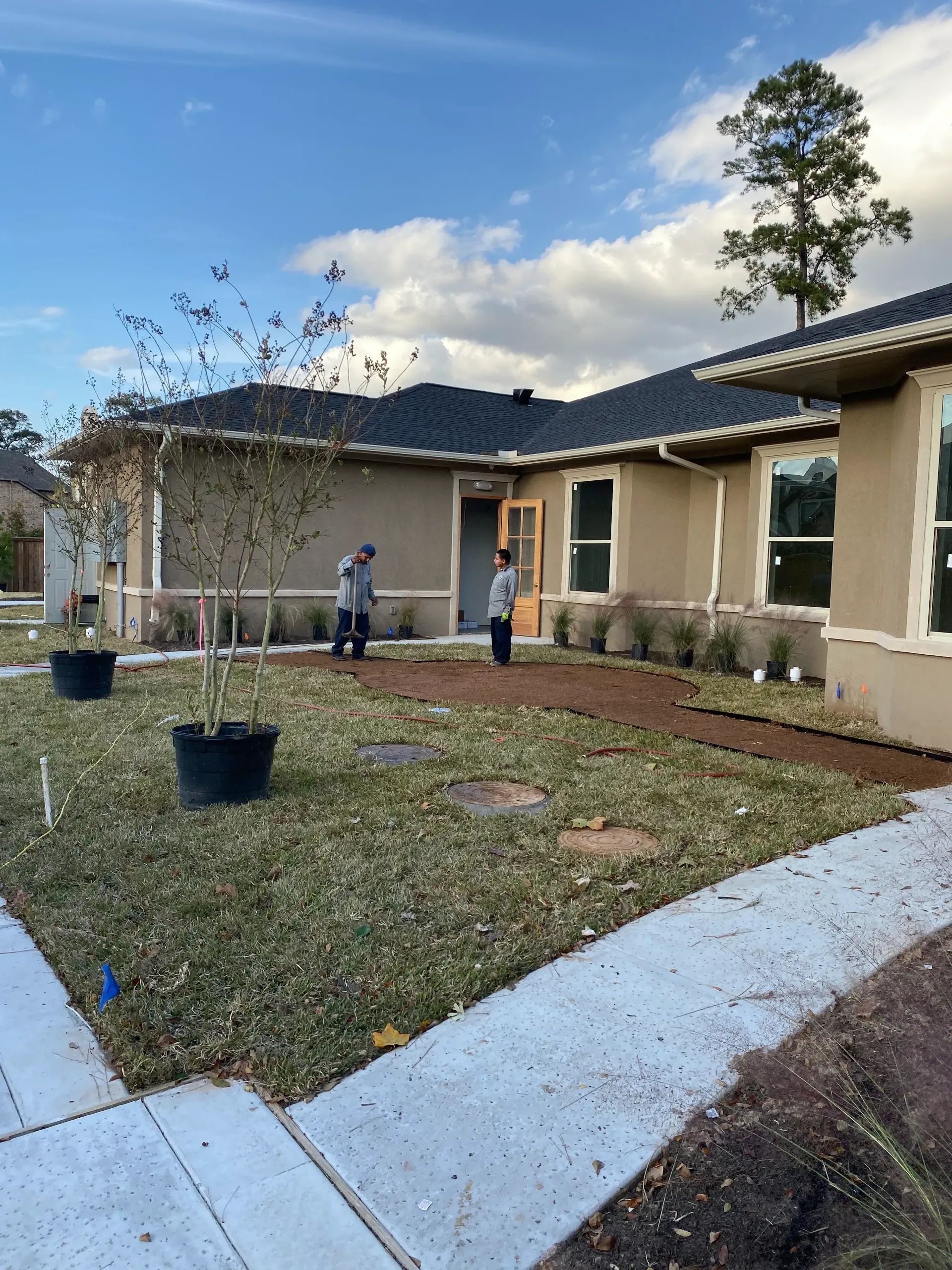 Backyard patio with two workers, potted plants, and a newly landscaped lawn beside a beige house.