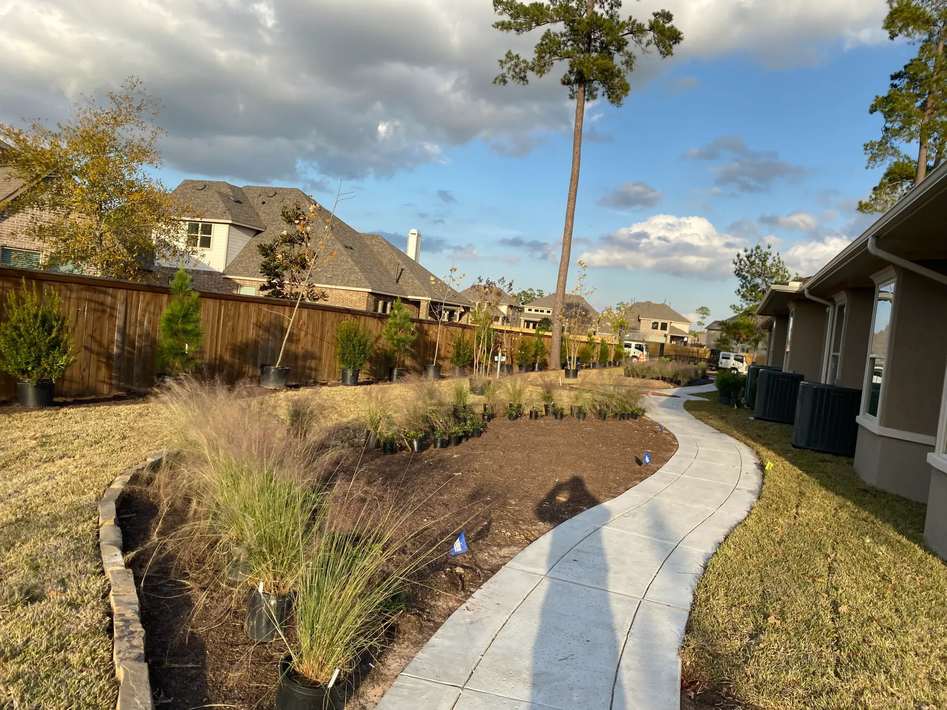 Curved garden path beside shrubs and houses under a partly cloudy sky, with long shadows.
