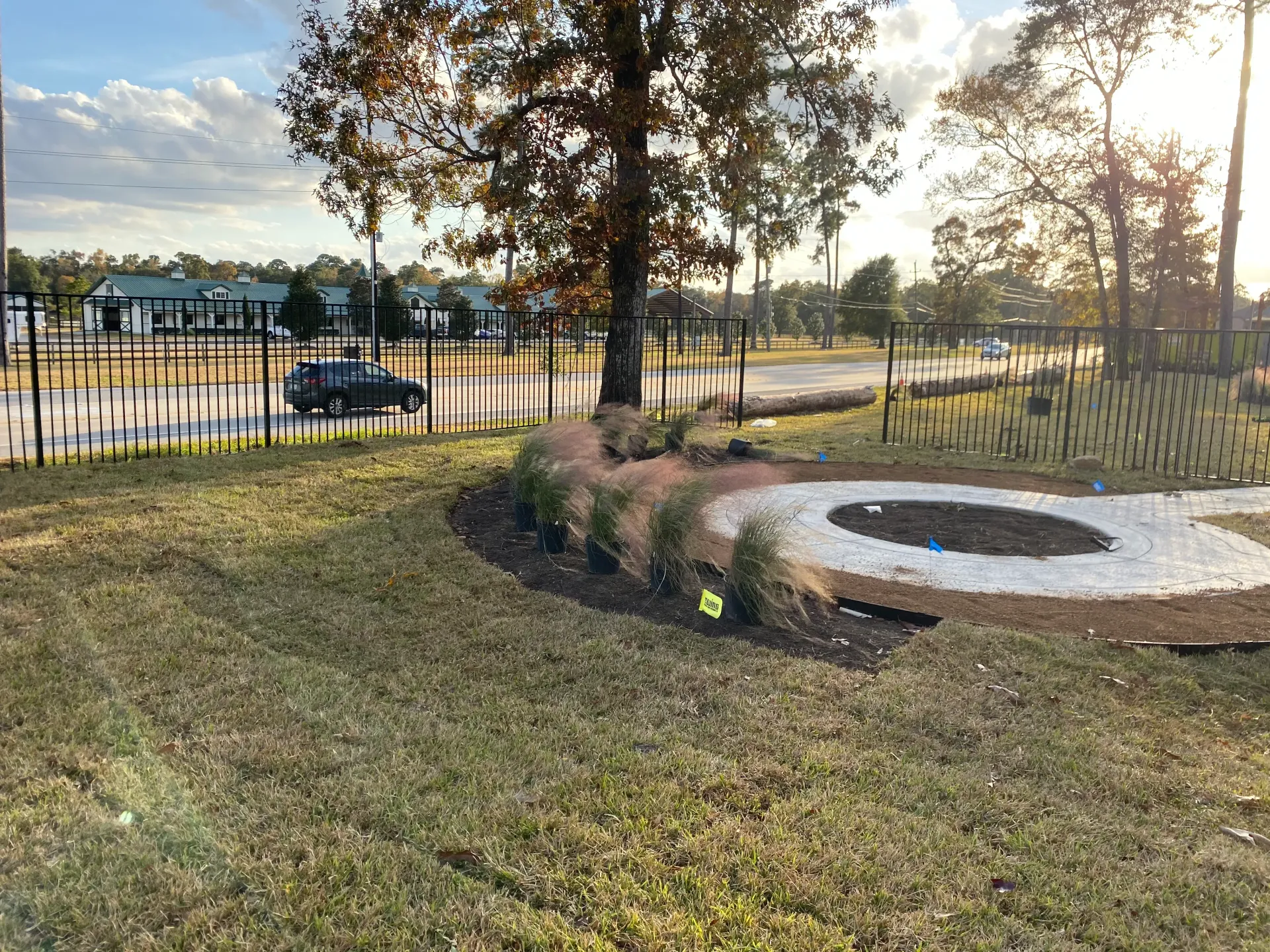 Sunlit park with a fenced pond, grassy field, trees, and a circular stone fire pit in the foreground