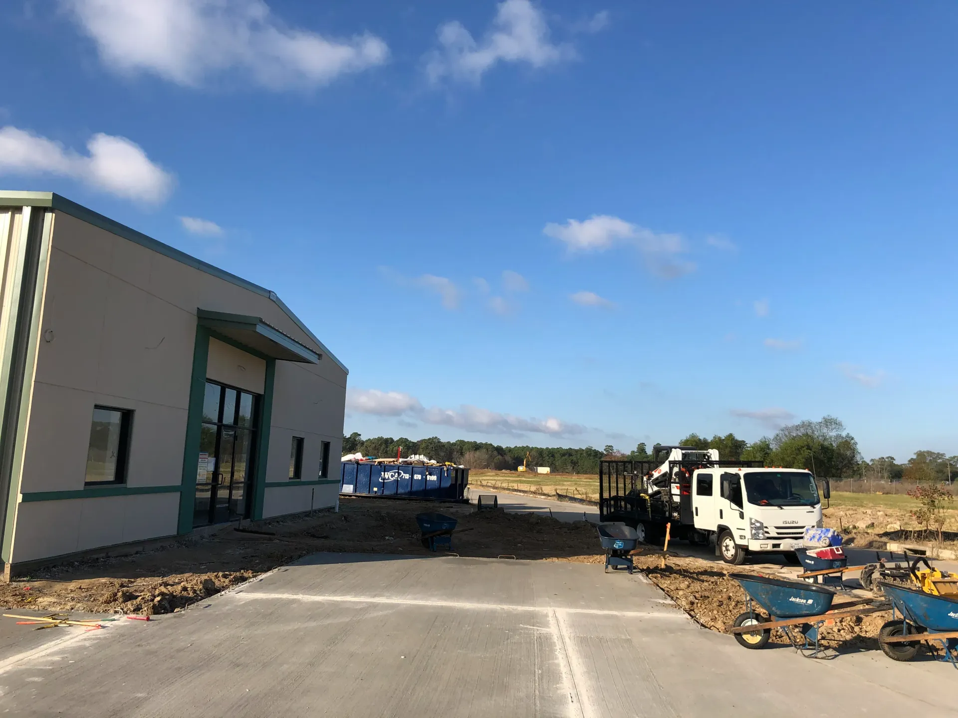 Construction site beside a white building, with a truck, equipment, and dirt under a blue sky.