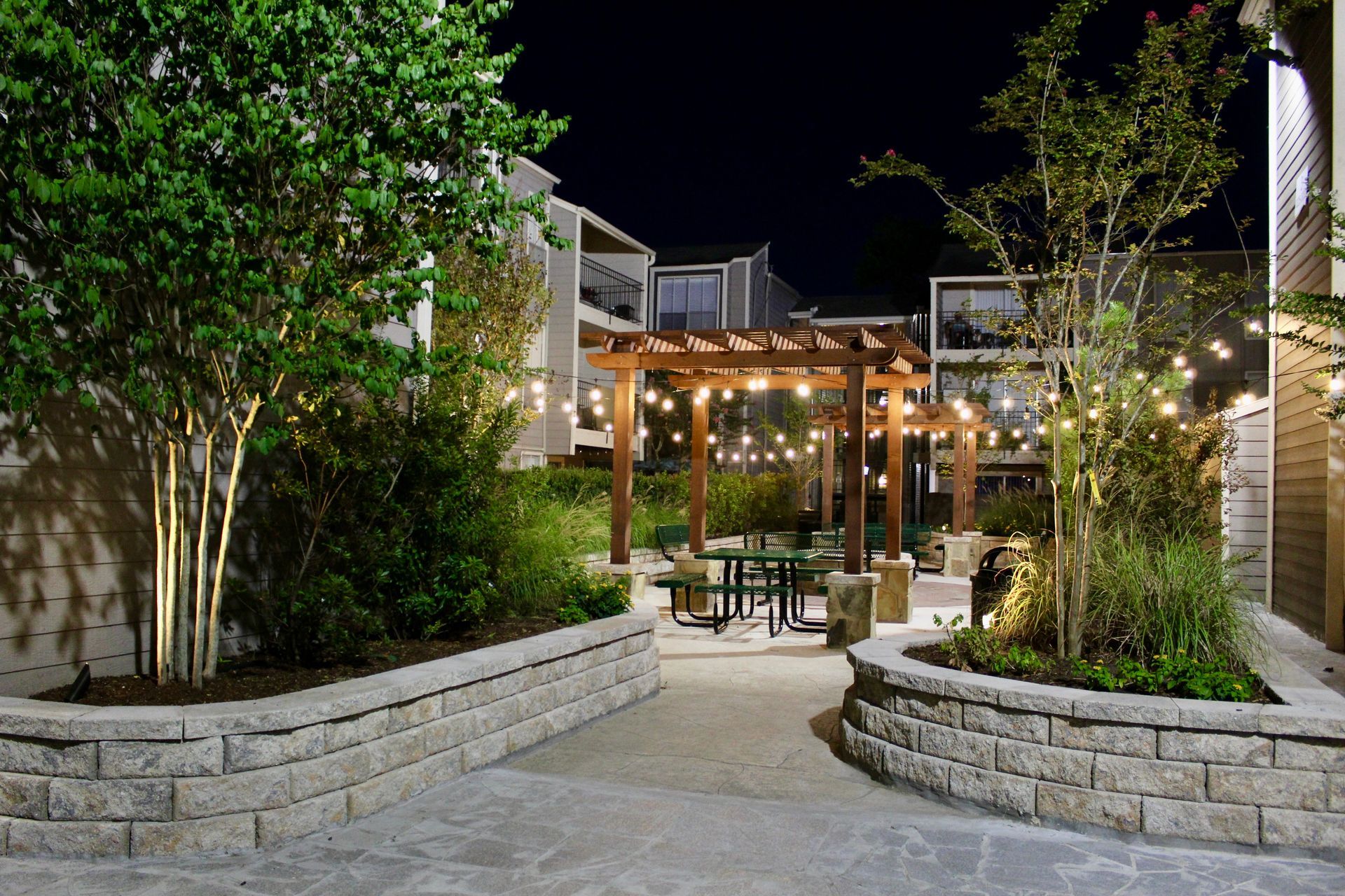 Nighttime courtyard dining area with string lights, wooden pergola, stone planters, and trees