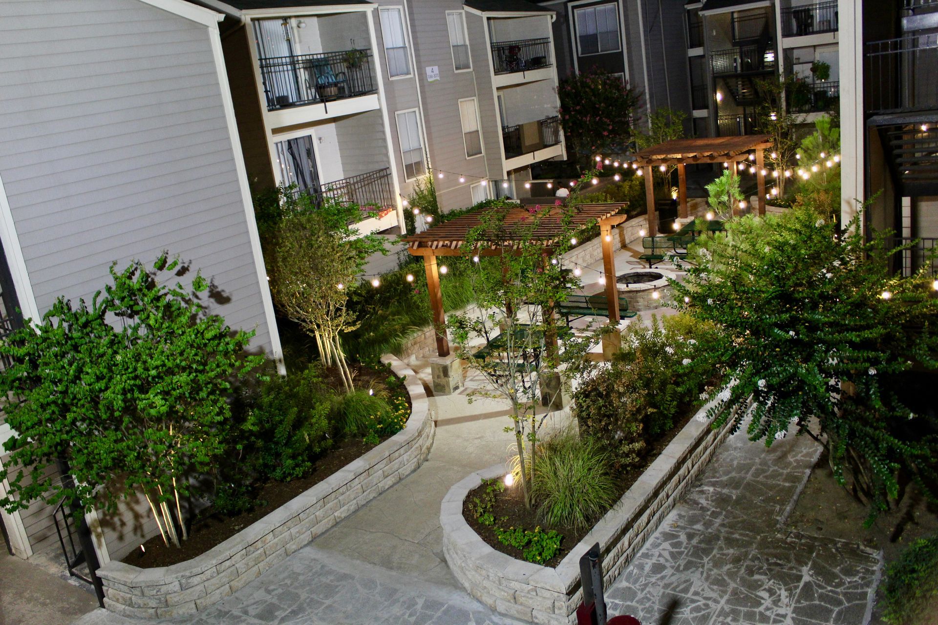 Outdoor courtyard with lit tables, trees, and walkway beside apartment buildings at dusk