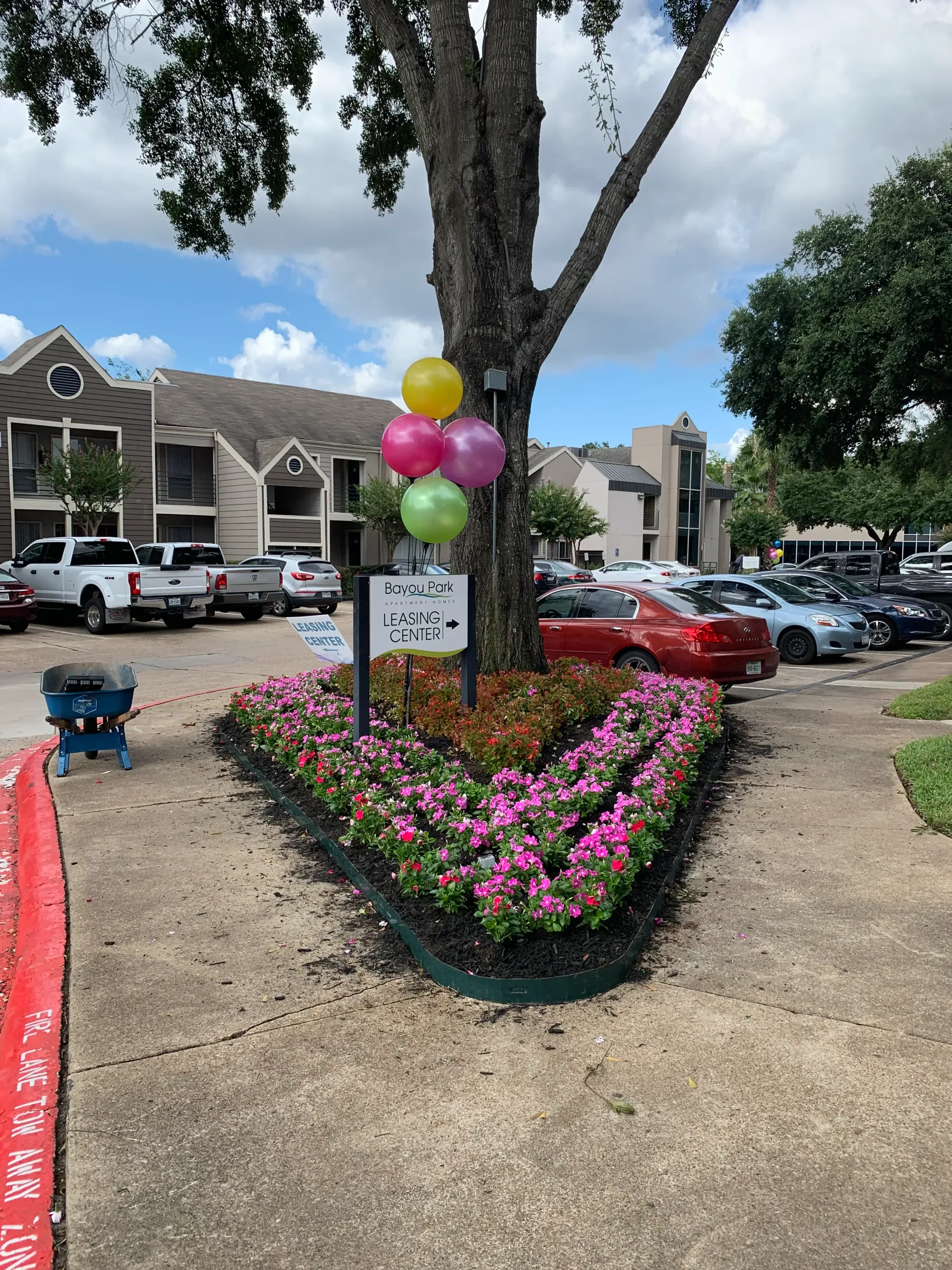 Balloon-decorated tree in a flower bed beside apartment parking lot with parked cars and cloudy sky