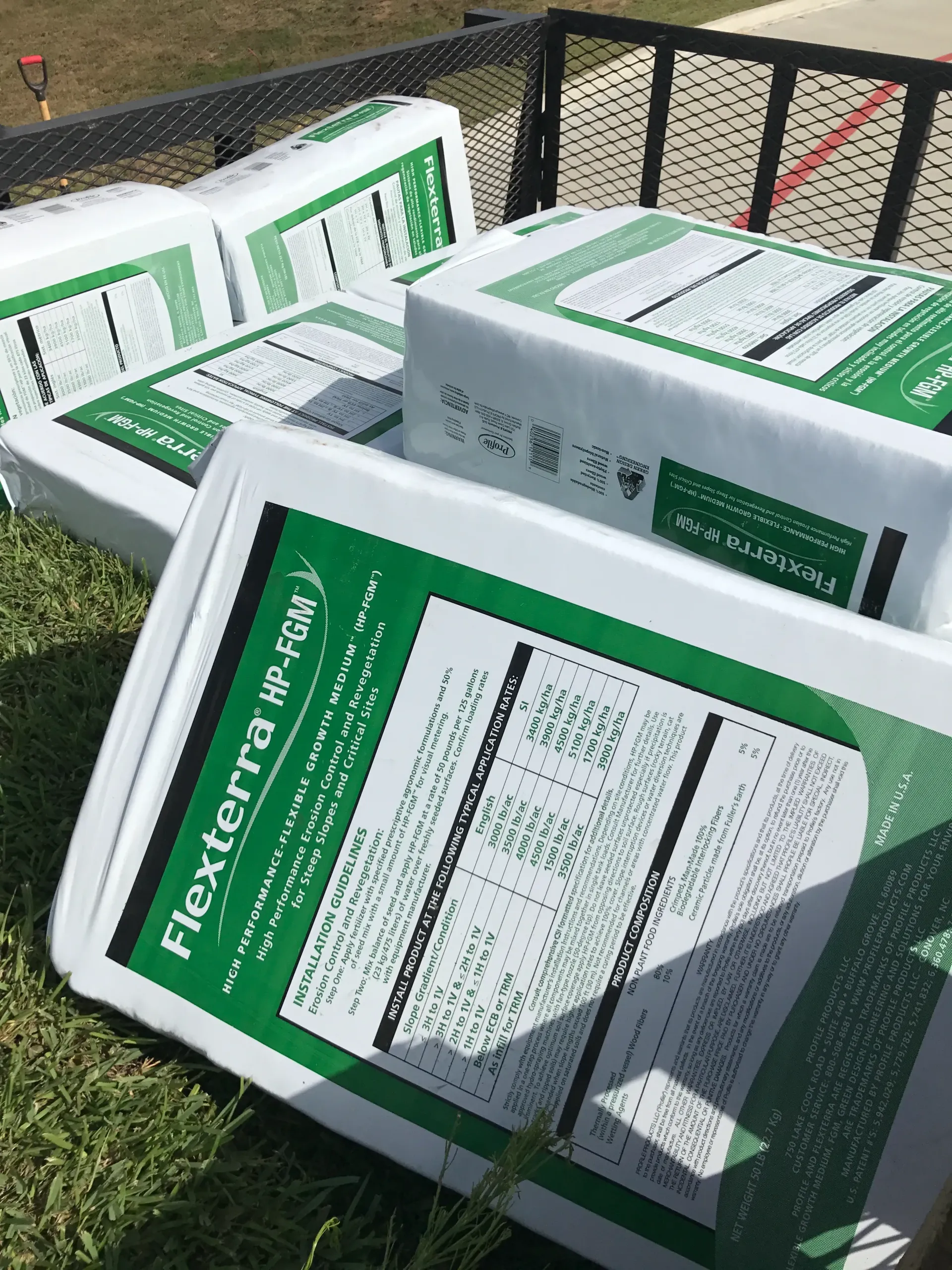 White and green plastic containers labeled “TREX” stacked outdoors on grass near a fence