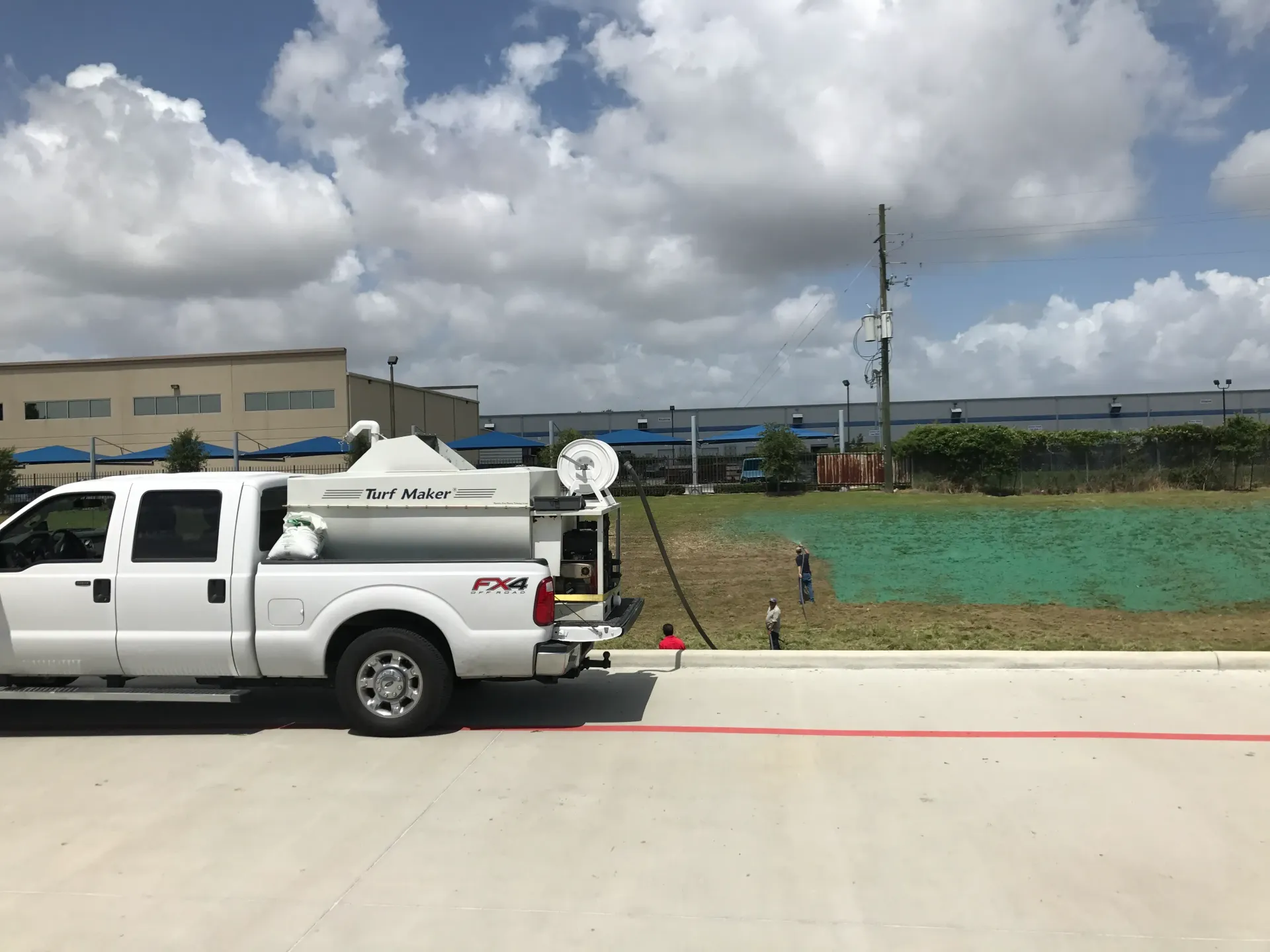 White utility truck parked by a curb beside a grassy lot and industrial buildings under a cloudy sky