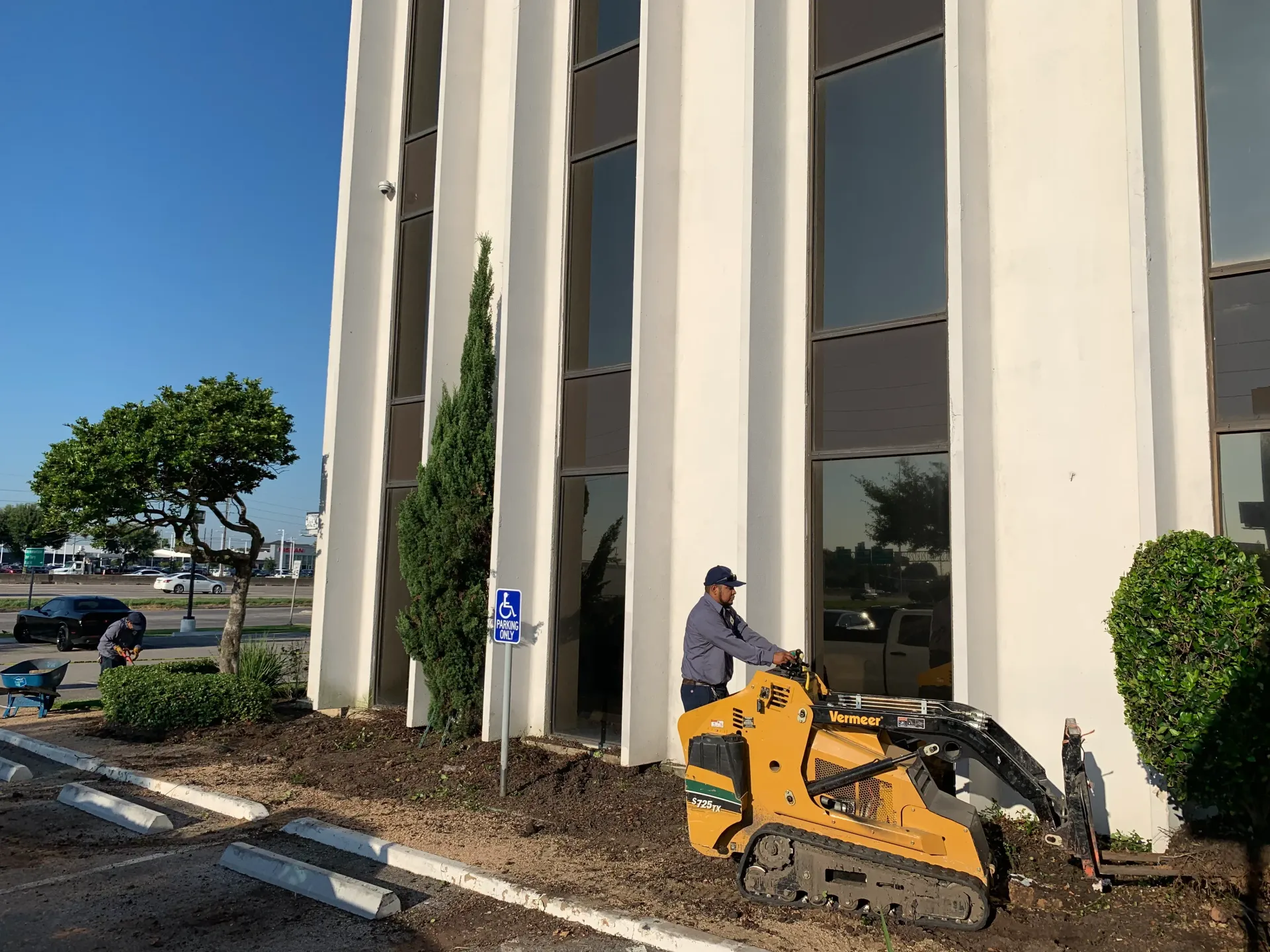 Worker on an orange lift near a tall beige building with trees and parked cars nearby