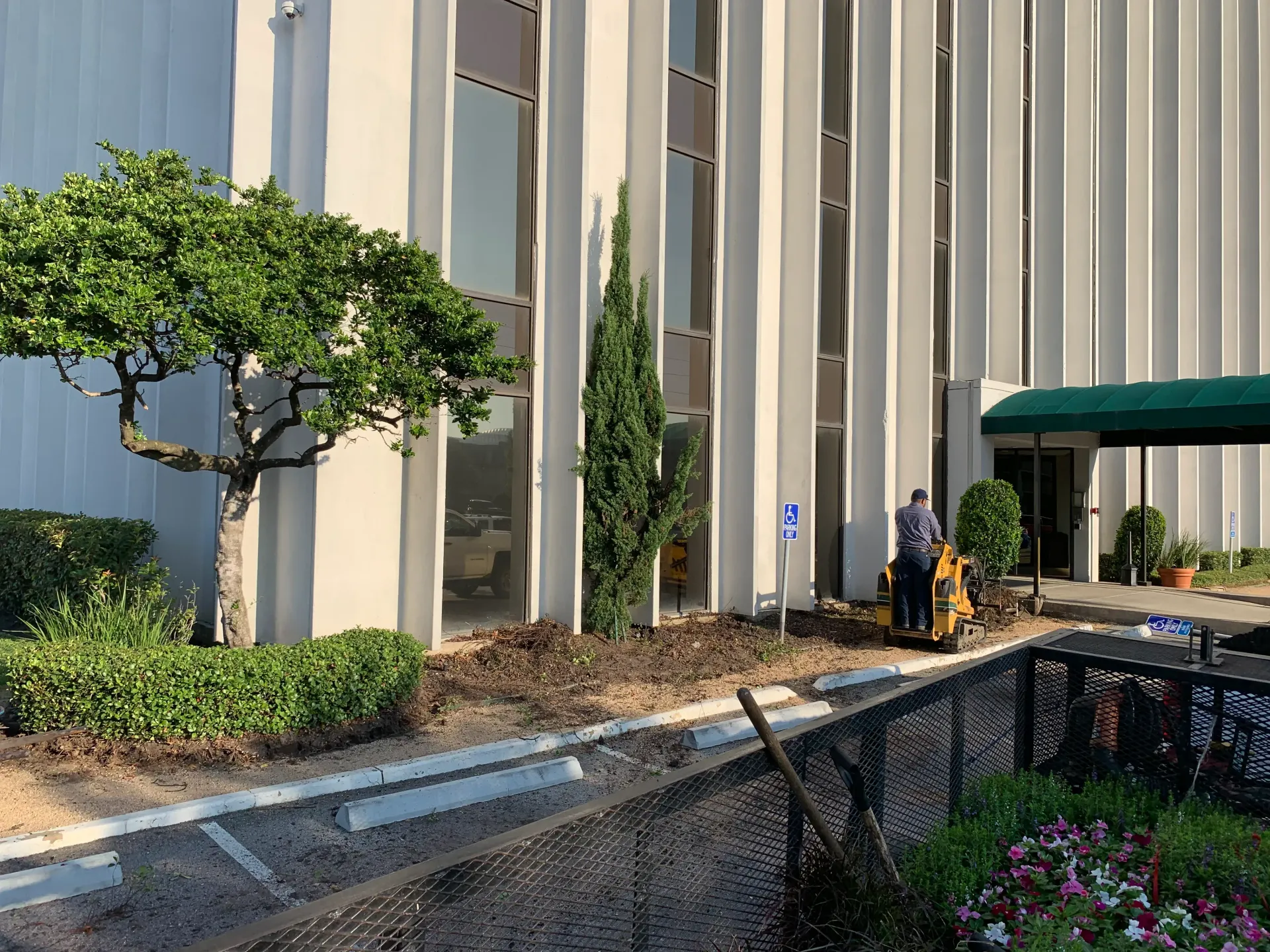 Building exterior with landscaping, a walkway, and two workers near the entrance under a green awning