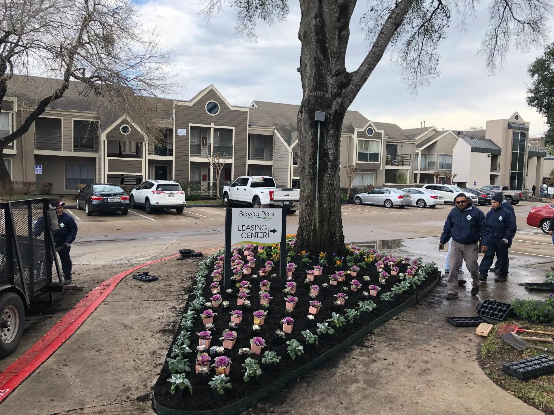 Memorial flower bed with a sign, two people walking nearby, and parked cars outside apartment buildings