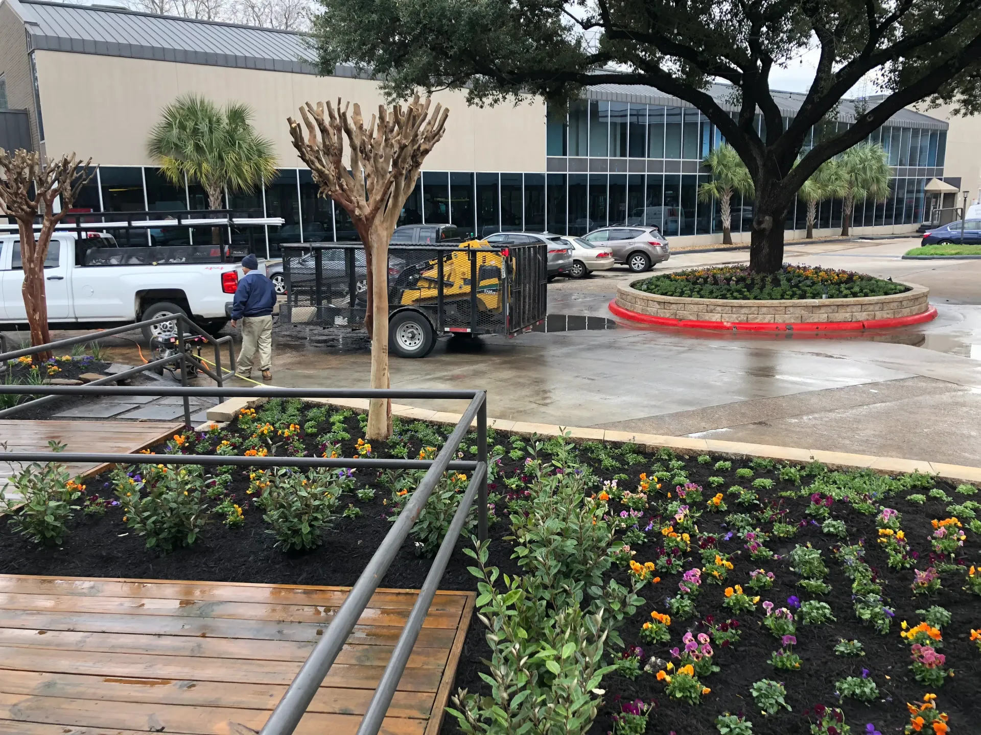Rainy parking lot with flower beds, parked cars, and a building in the background.