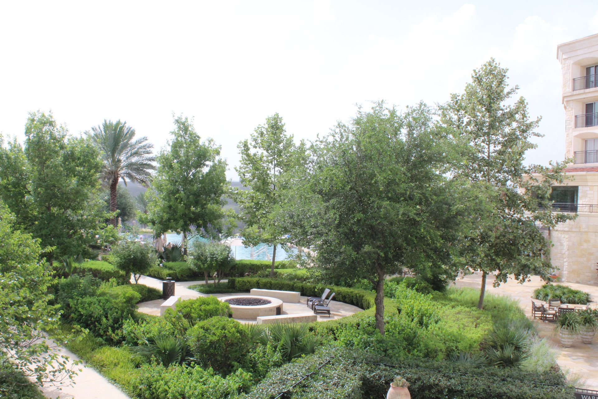 Lush garden courtyard with trees, shrubs, stone paths, and a small seating area beside a building