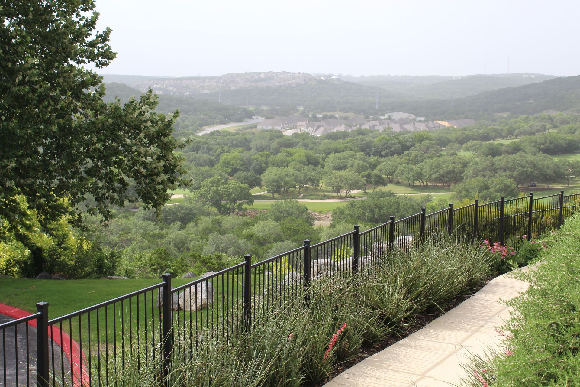 Terraced garden path overlooking a green valley and hazy hills, with a black fence and flowers in the foreground