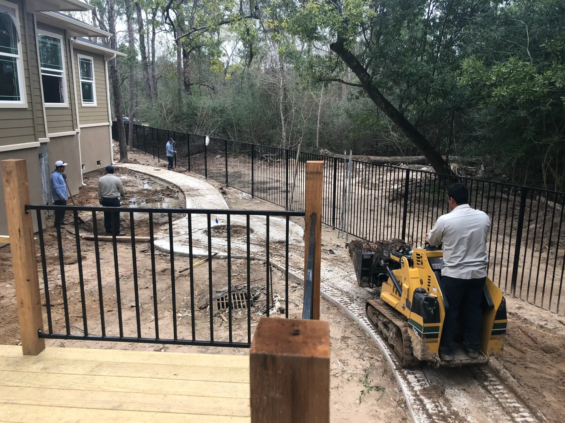 Person on a yellow compacting machine smoothing dirt beside a black fence and house in a yard