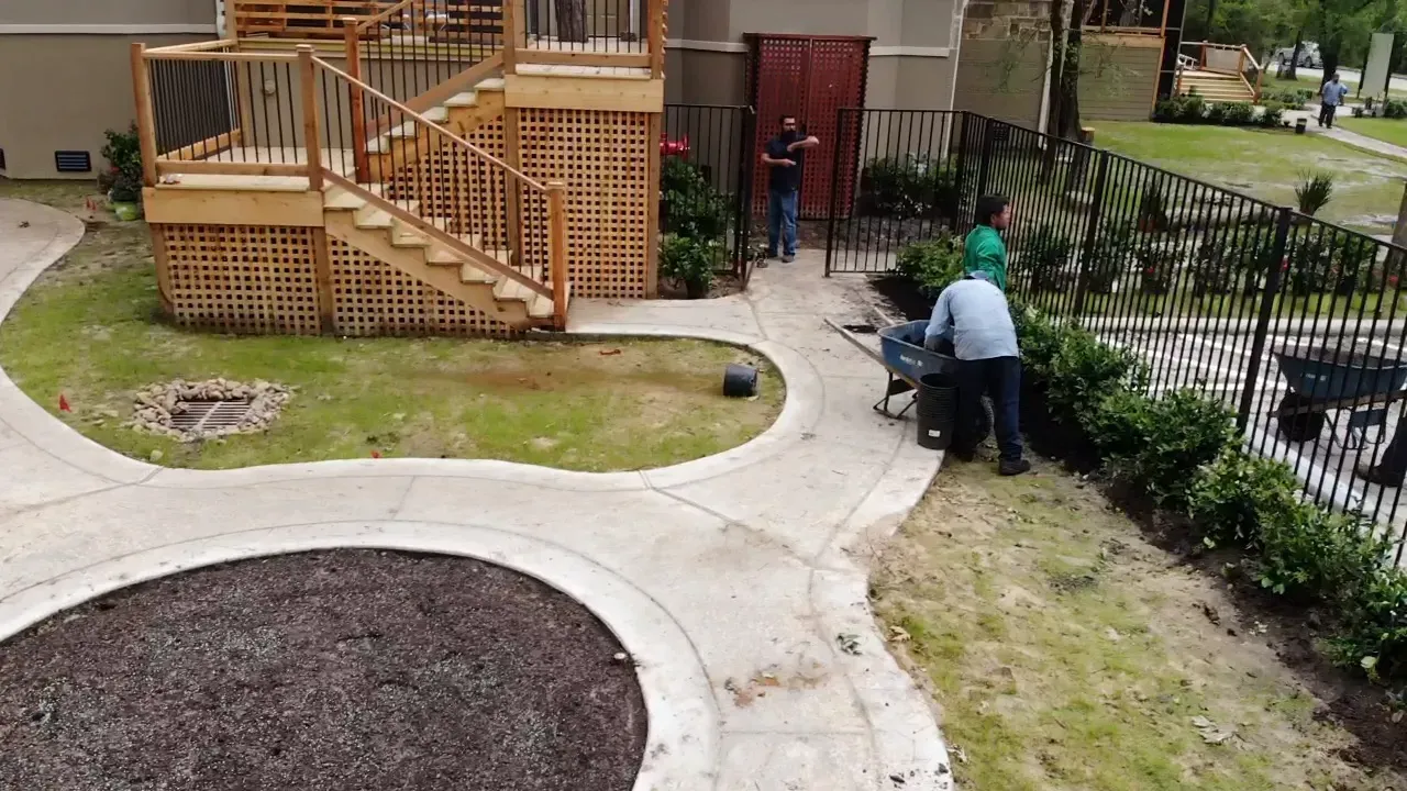 Person pushing a mower on a residential sidewalk beside a wooden deck and landscaped yard.