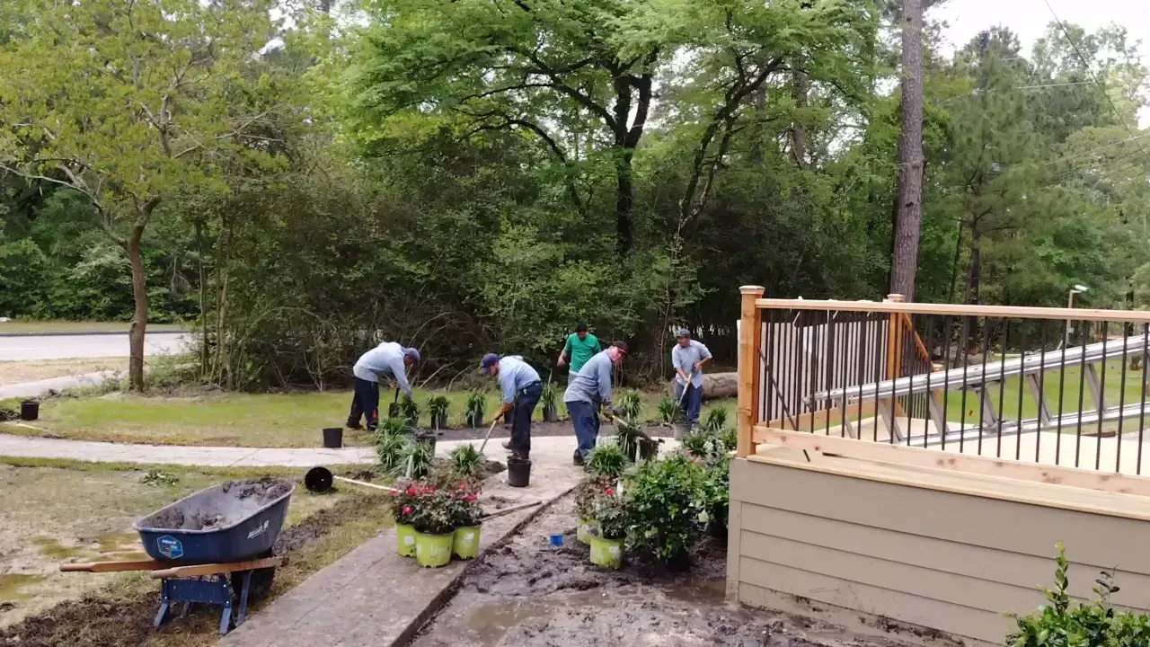 Workers planting flowers along a wooded path beside a house porch