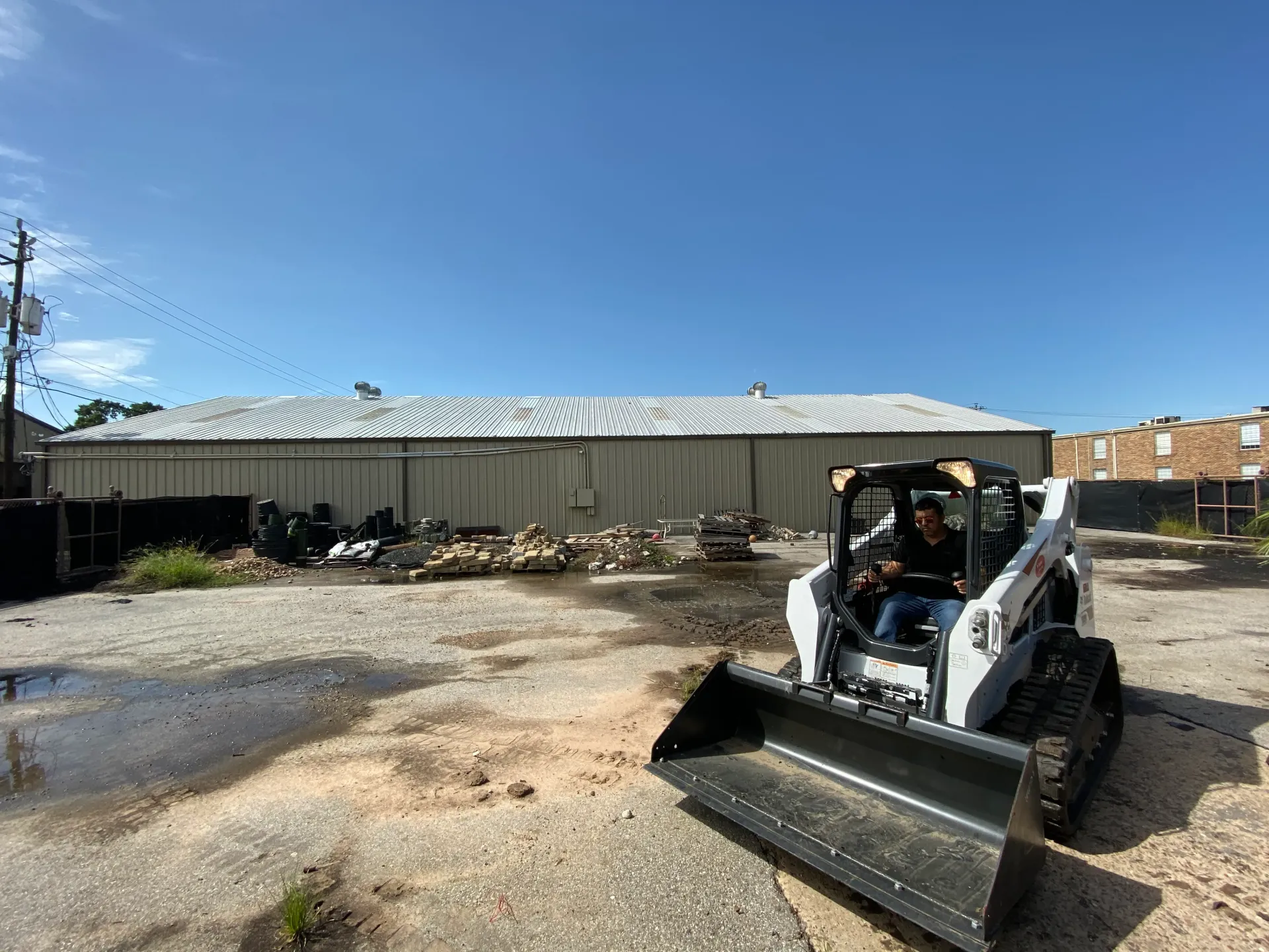 White skid steer with bucket on gravel lot beside a corrugated fence under blue sky