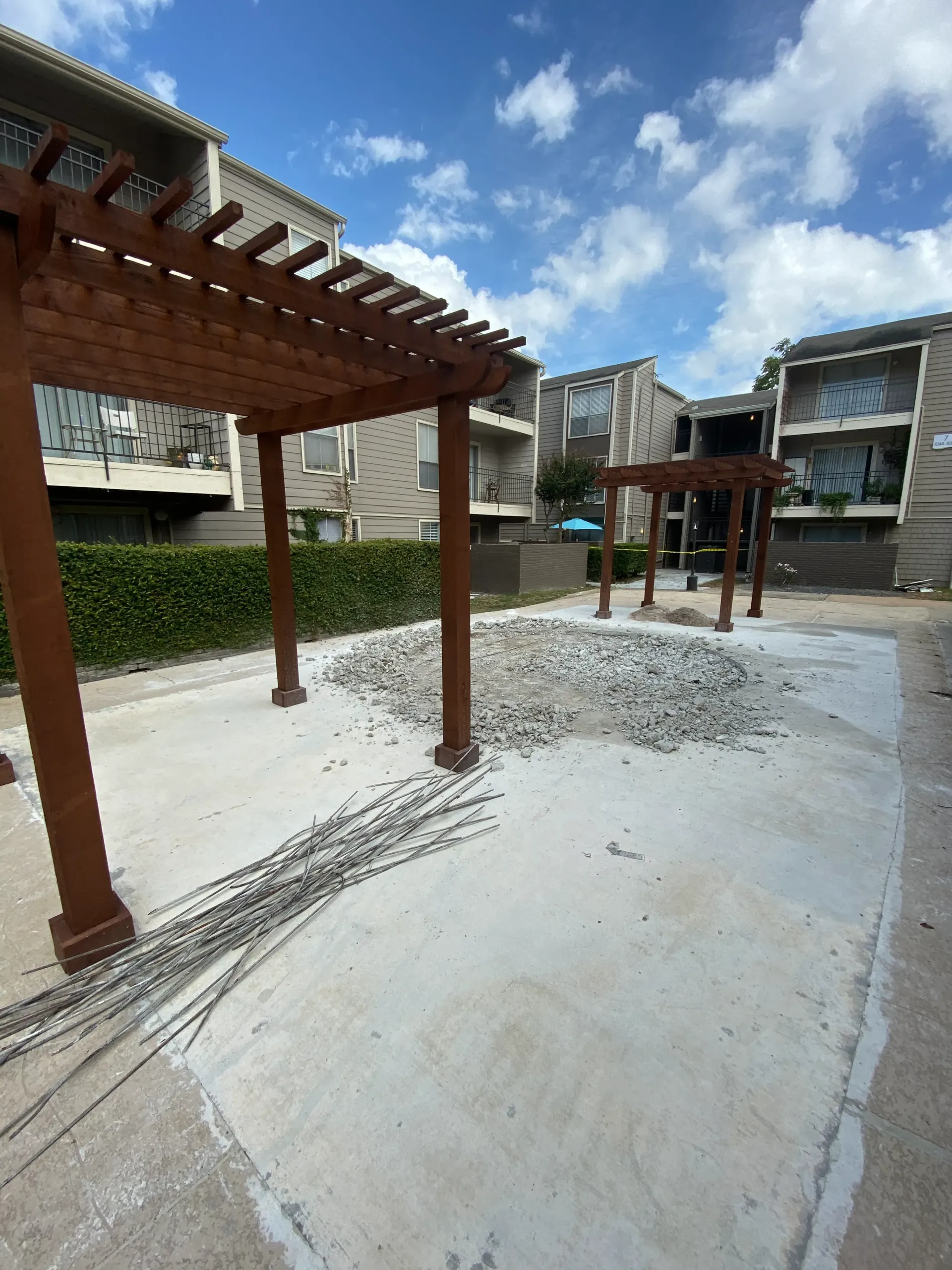 Outdoor courtyard with wooden pergolas, gravel paths, and apartment buildings under a partly cloudy sky