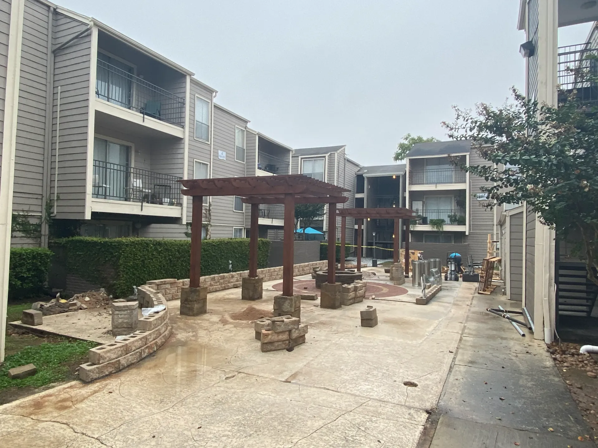 Apartment walkway under construction with wooden pergolas, scattered bricks, and workers in the distance