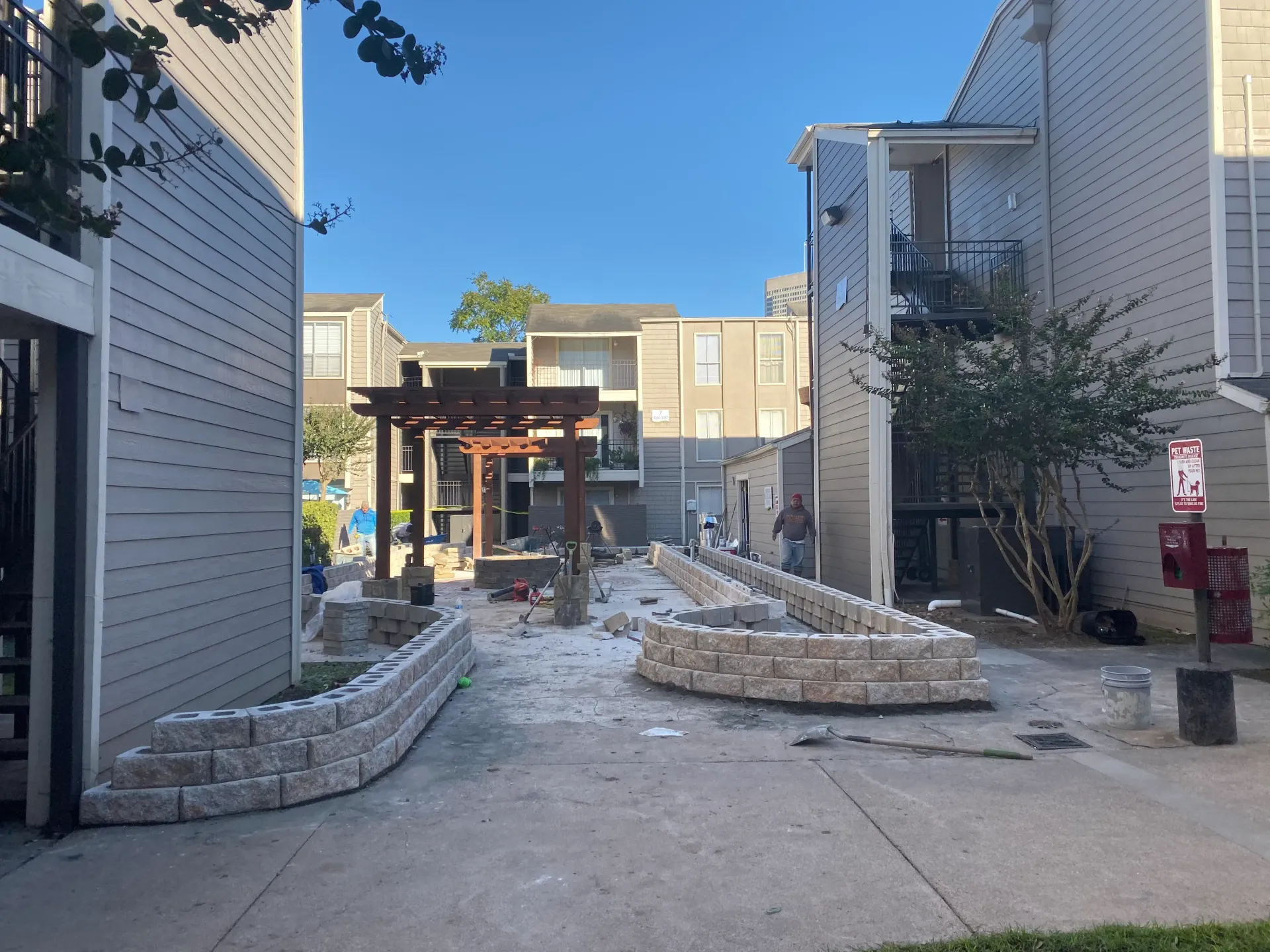 Narrow outdoor walkway between buildings with stone planters, a small pergola, and blue sky overhead