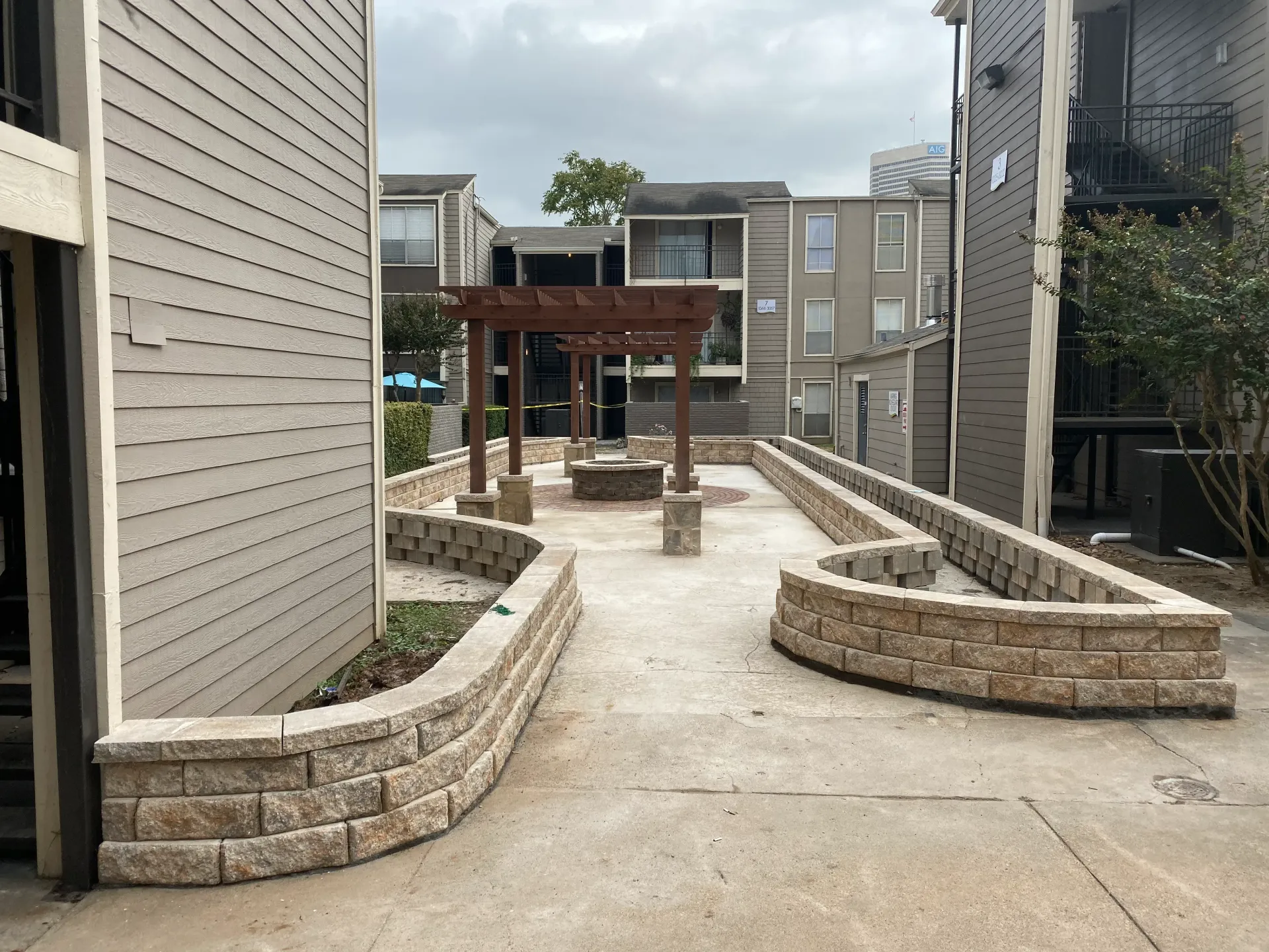 Concrete courtyard path with low stone planters leading to a wooden pergola between apartment buildings