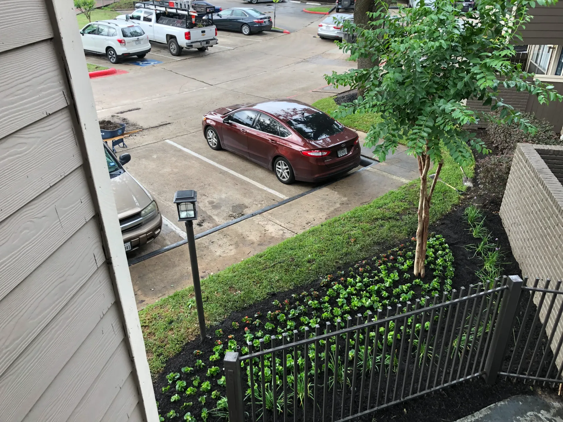 Parking lot seen from an apartment balcony with a maroon SUV, black fence, trees, and parked cars in the background