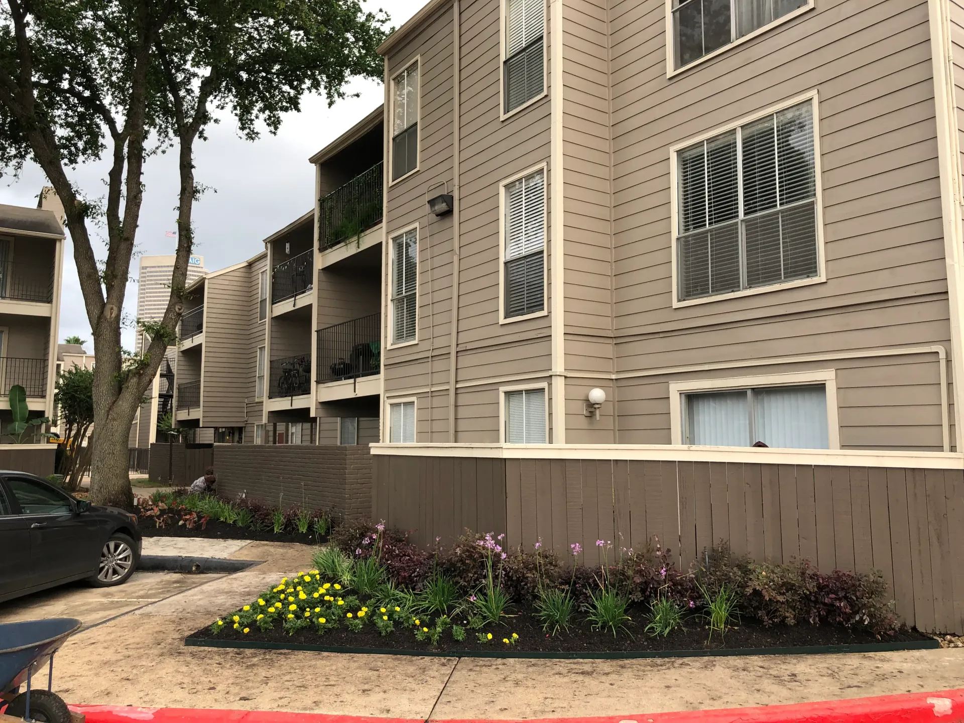 Apartment building exterior with beige siding, trees, and landscaped flower beds along a walkway