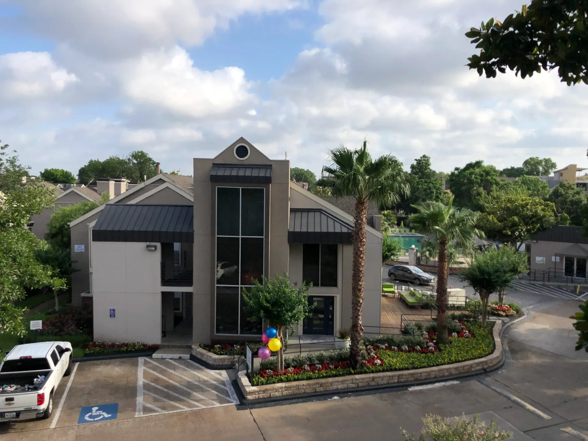 Modern two-story building with glass entrance, landscaped grounds, and parked cars under a partly cloudy sky