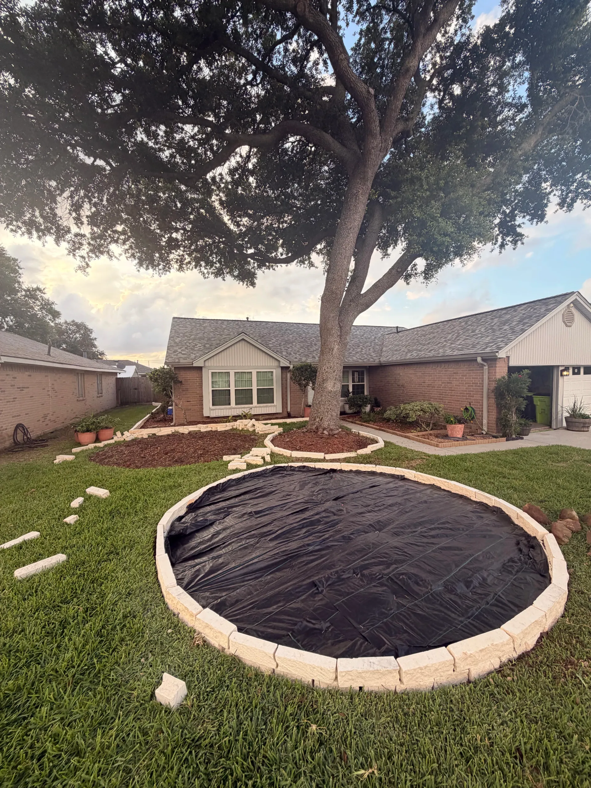 Backyard with a large tree, stone patio, and a round covered garden bed surrounded by grass.