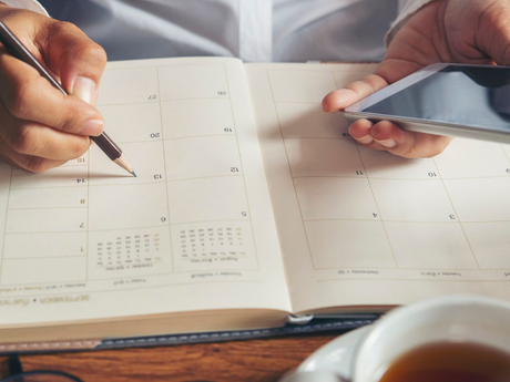 A person holds a smartphone in one hand and uses a pencil to write in a paper planner at a desk.